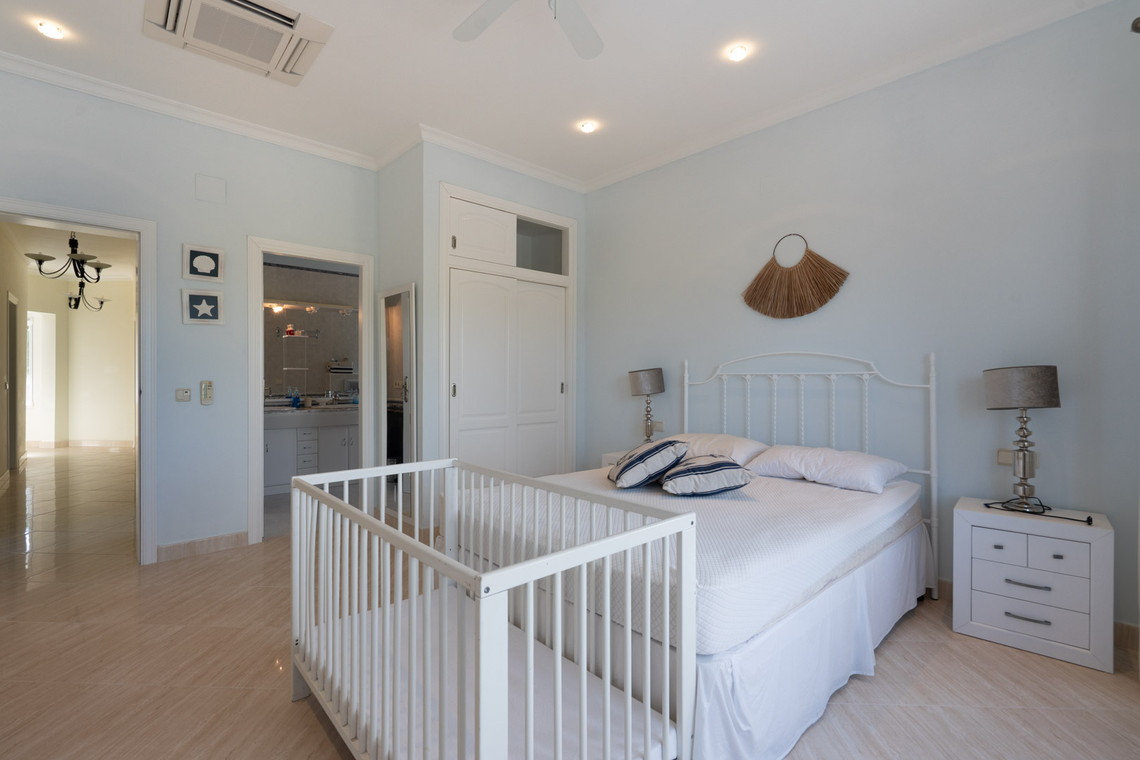 Chambre lumineuse aux murs bleu pâle avec lit double, tête de lit en fer forgé blanc, berceau, placards intégrés et accès à une salle de bain attenante.
