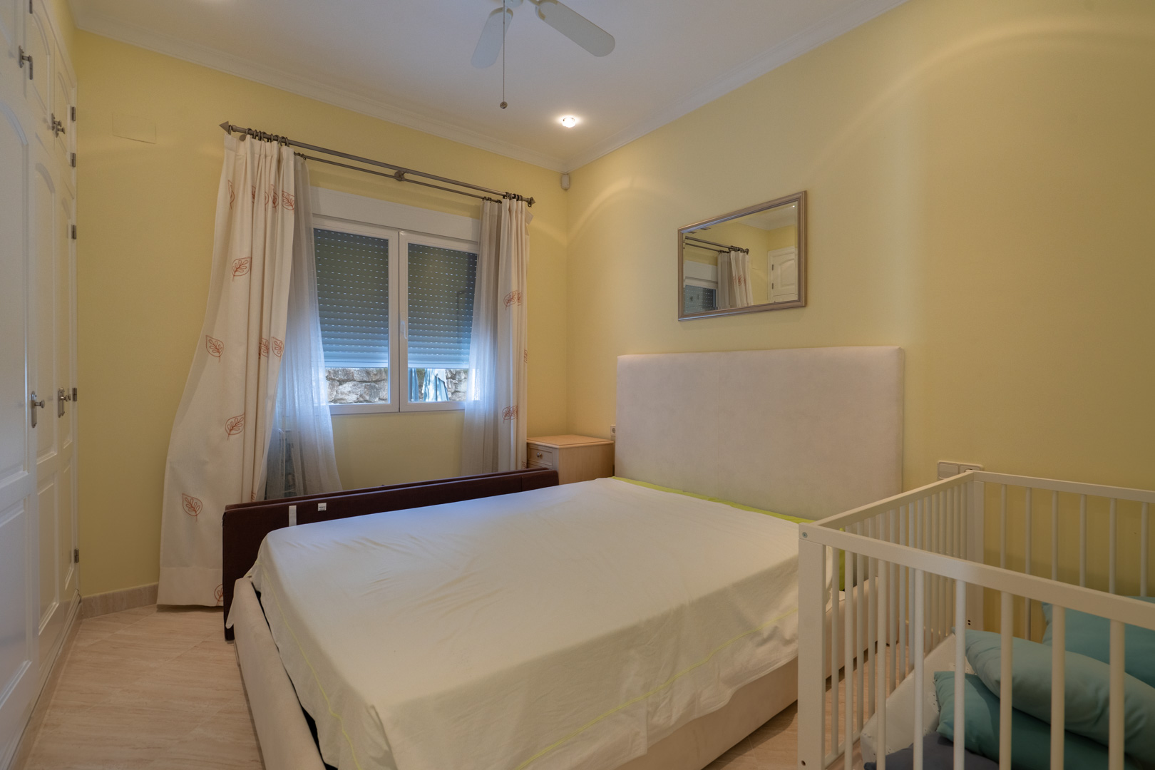 Chambre lumineuse aux tons crème avec placards intégrés blancs, lit double à tête de lit rembourrée et berceau. Comprend un ventilateur de plafond et un éclairage encastré.