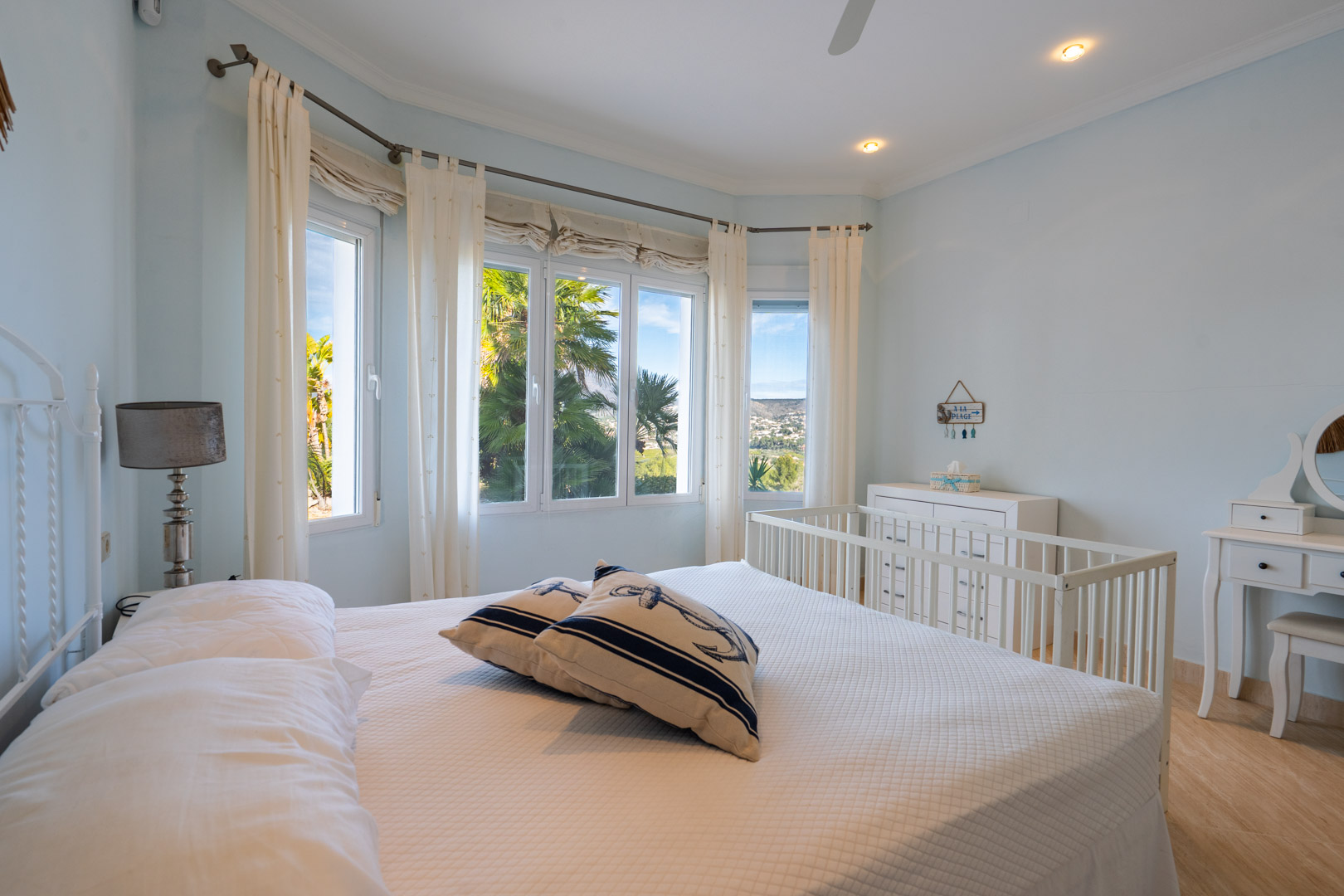 Chambre lumineuse avec baie vitrée offrant une vue sur le Montgó, mobilier blanc comprenant un berceau et une coiffeuse, et décoration nautique.
