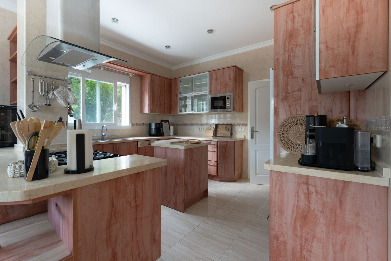 Cuisine équipée avec meubles en bois rougeâtre, plans de travail en pierre claire, îlot central et hotte aspirante en verre sur plaque de cuisson au gaz.
