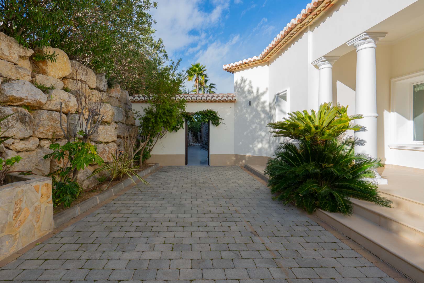 Entrée extérieure avec allée pavée en gris, mur de soutènement en pierre naturelle et façade blanche agrémentée de colonnes classiques.