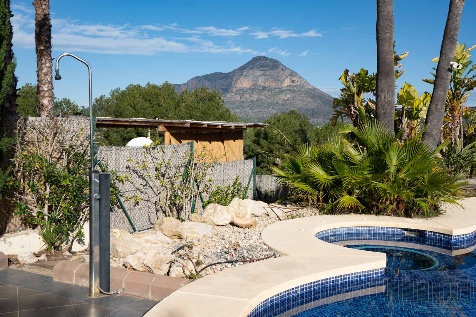 Espace piscine avec douche extérieure en inox, entouré d'une végétation tropicale offrant une vue dégagée sur le mont Montgó.