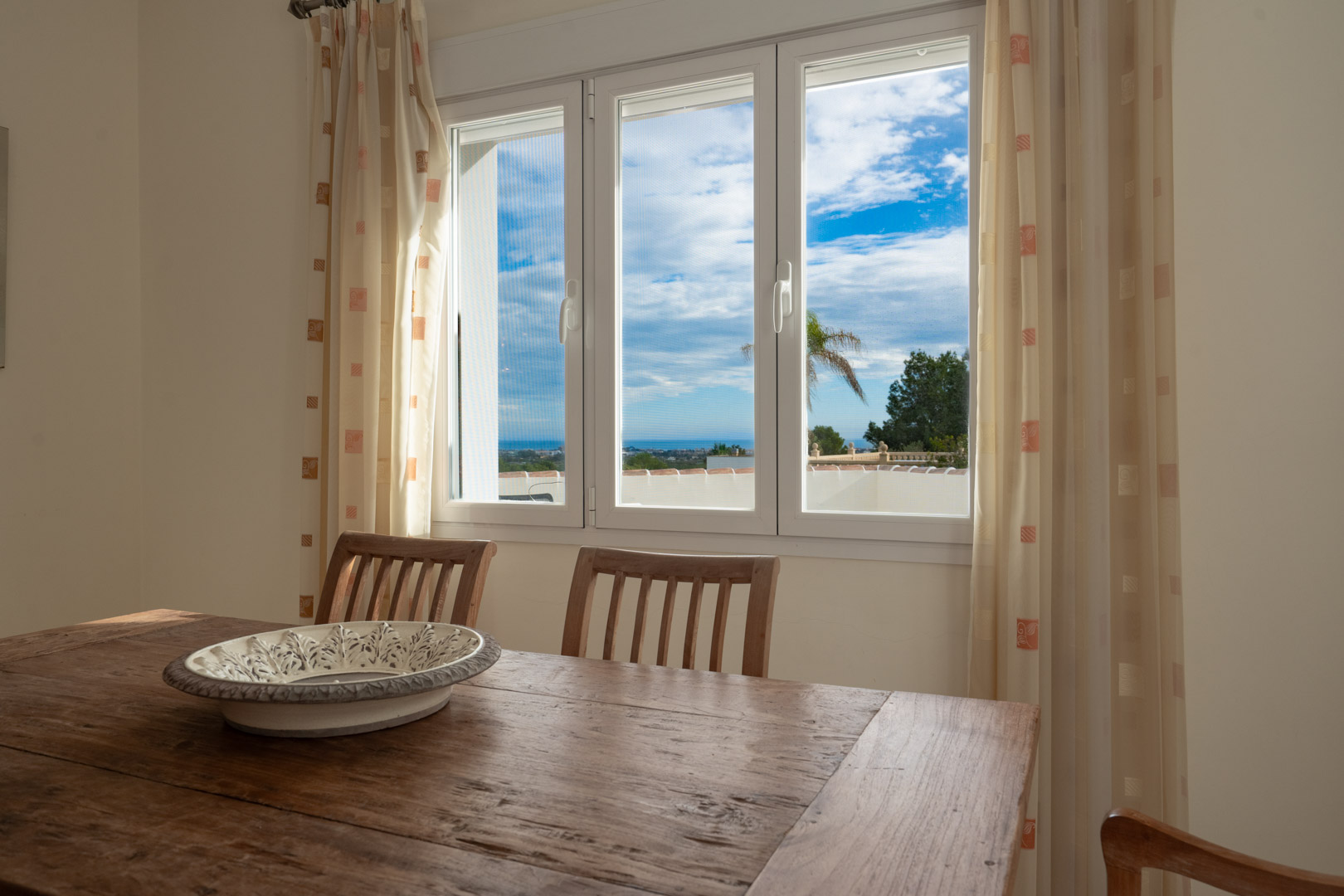 Espace repas avec table en bois massif offrant une vue sur la mer Méditerranée à travers une fenêtre blanche à trois vantaux baignée de lumière.