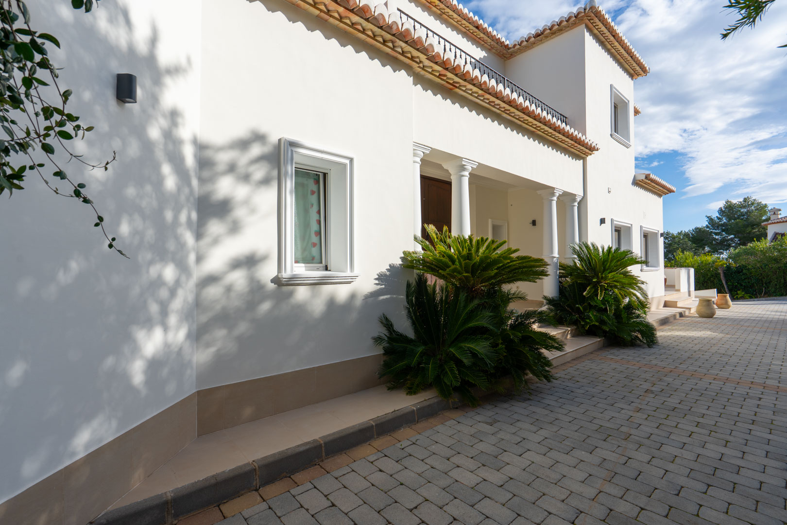 Façade méditerranéenne avec colonnes blanches, allée pavée et détails architecturaux incluant des tuiles en terre cuite et une végétation soignée.
