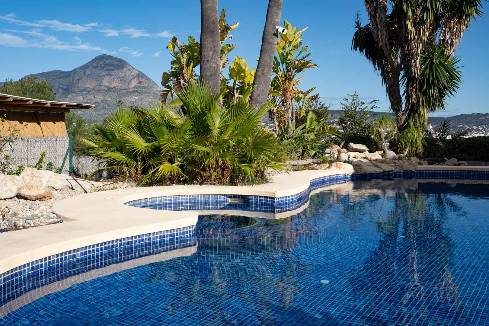 Piscine aux formes arrondies en mosaïque bleue avec jacuzzi intégré, entourée d'un jardin tropical offrant une vue dégagée sur le mont Montgó.