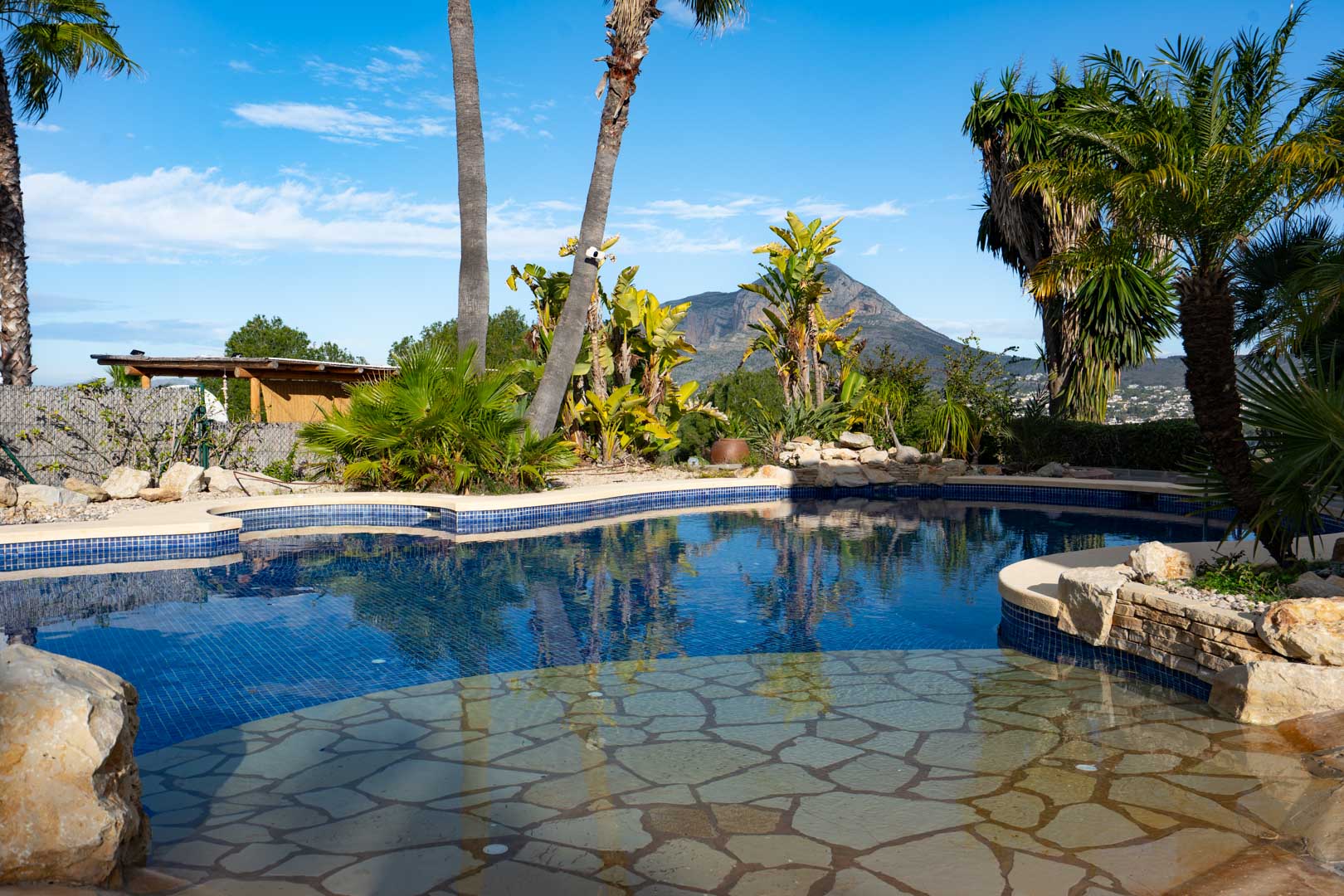 Piscine aux formes organiques avec mosaïque bleue et entrée en pierre naturelle, entourée d'un jardin tropical offrant une vue sur le mont Montgó.