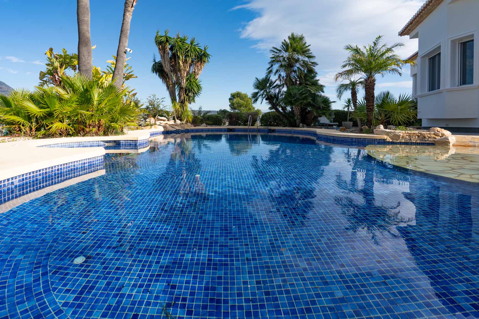 Piscine en mosaïque bleue au design irrégulier, entourée d'un jardin tropical avec des palmiers et une végétation méditerranéenne.