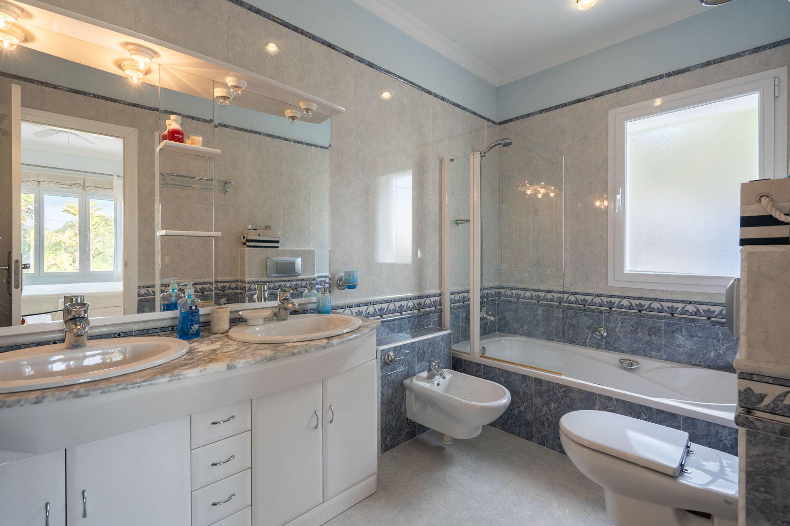 Salle de bain complète avec double vasque sur plan en marbre, baignoire avec paroi vitrée et carrelage élégant aux tons bleus et gris avec frises.
