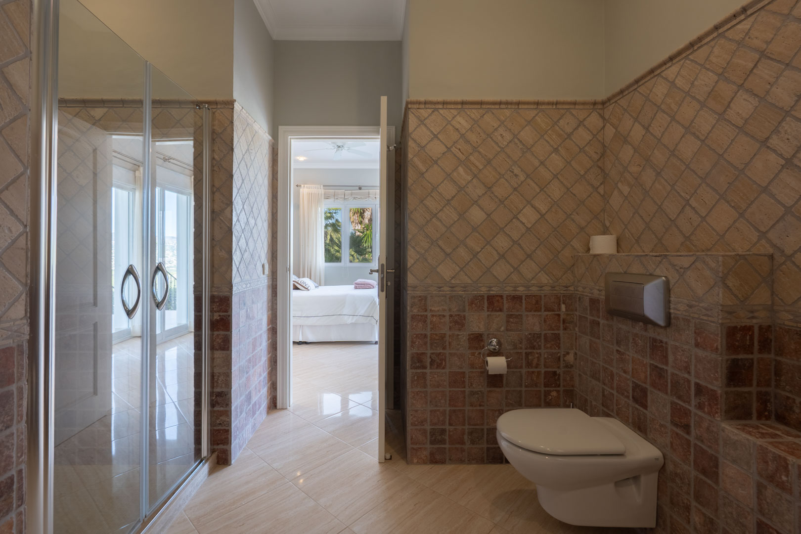 Salle de bains attenante avec carrelage rustique, toilettes suspendues et vue sur la chambre principale à travers la porte ouverte.