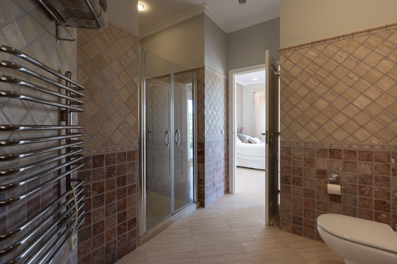 Salle de bains attenante avec douche vitrée, sèche-serviettes chromé et carrelage décoratif aux tons terreux, ouvrant sur une chambre lumineuse.