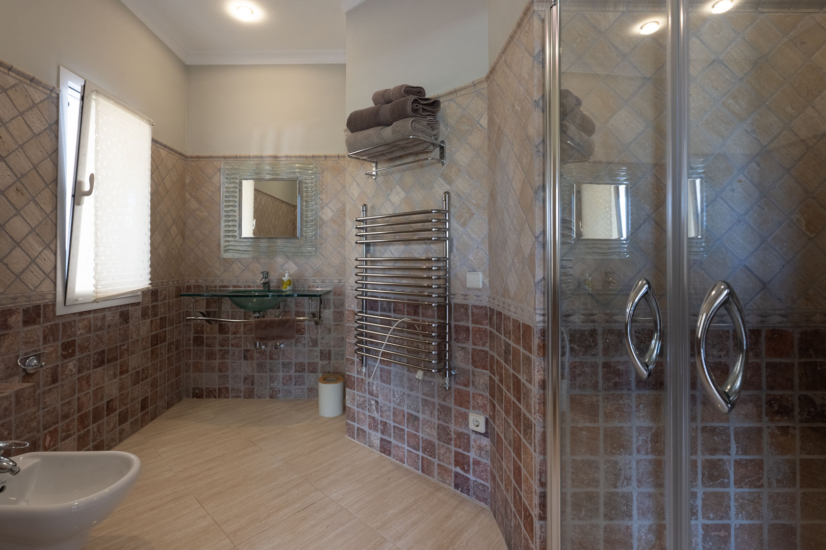 Salle de bains avec carrelage mural aux tons terreux, vasque en verre, radiateur sèche-serviettes chromé et cabine de douche vitrée.
