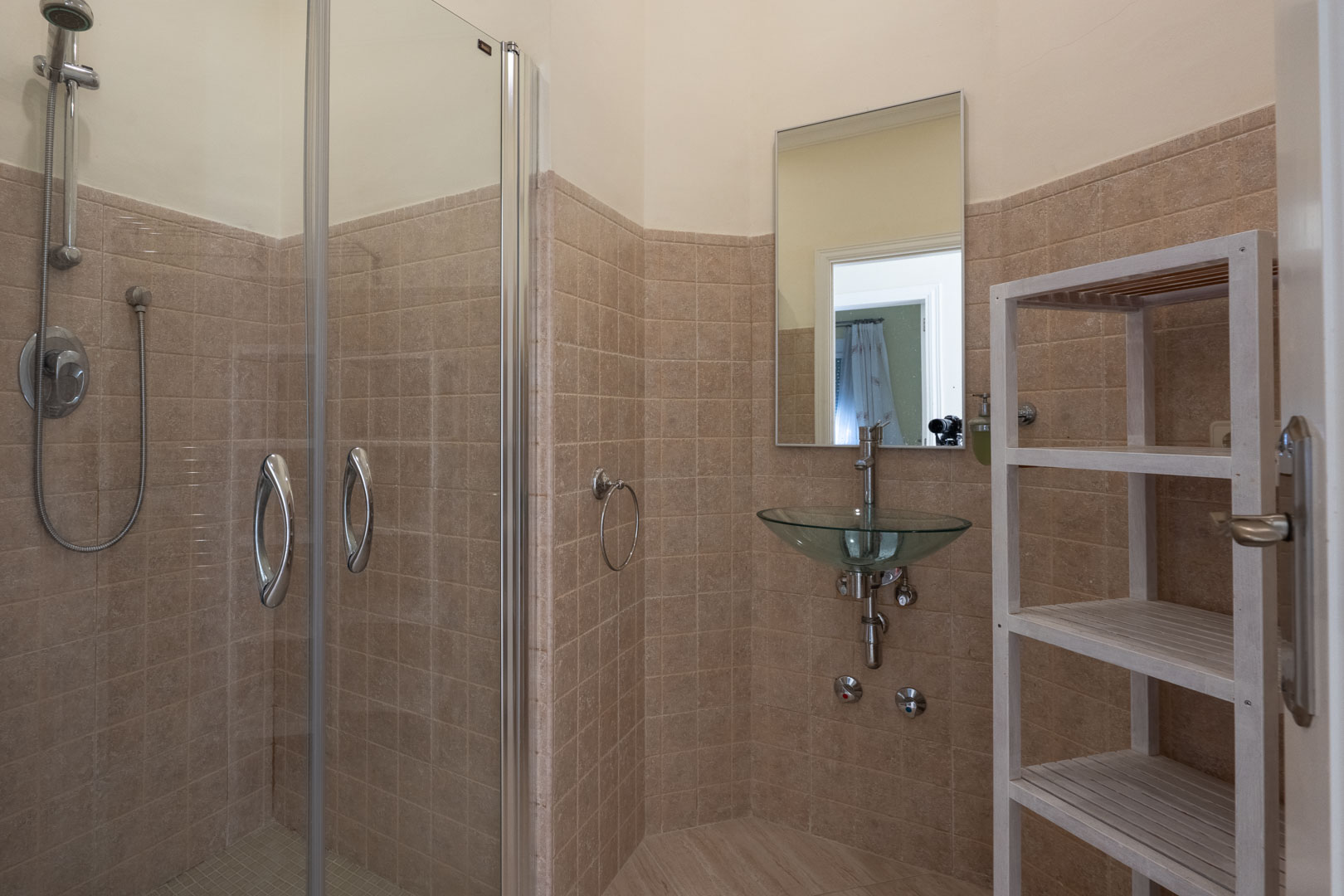 Salle d'eau comprenant une douche vitrée, un lavabo vasque en verre suspendu, des murs carrelés dans les tons beiges et une étagère de rangement en bois blanc.