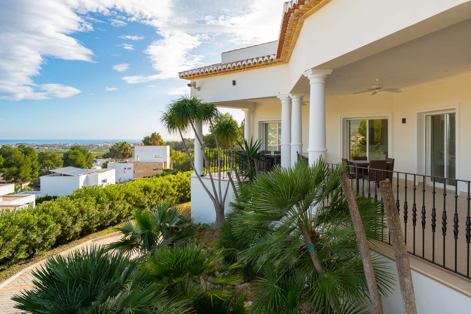 Terrasse couverte avec colonnes classiques et garde-corps en fer forgé, entourée de palmiers tropicaux avec une vue panoramique sur la mer Méditerranée.