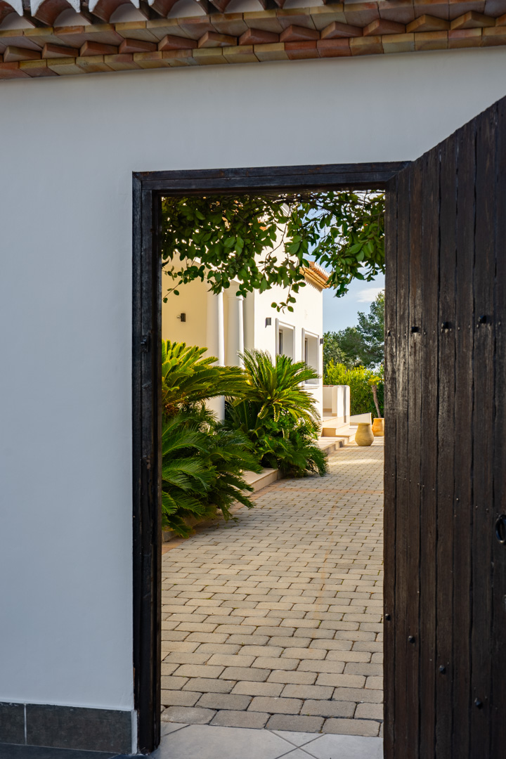 Vue à travers une porte en bois sombre ouvrant sur une allée pavée, bordée de palmiers et de la façade blanche immaculée de la propriété.