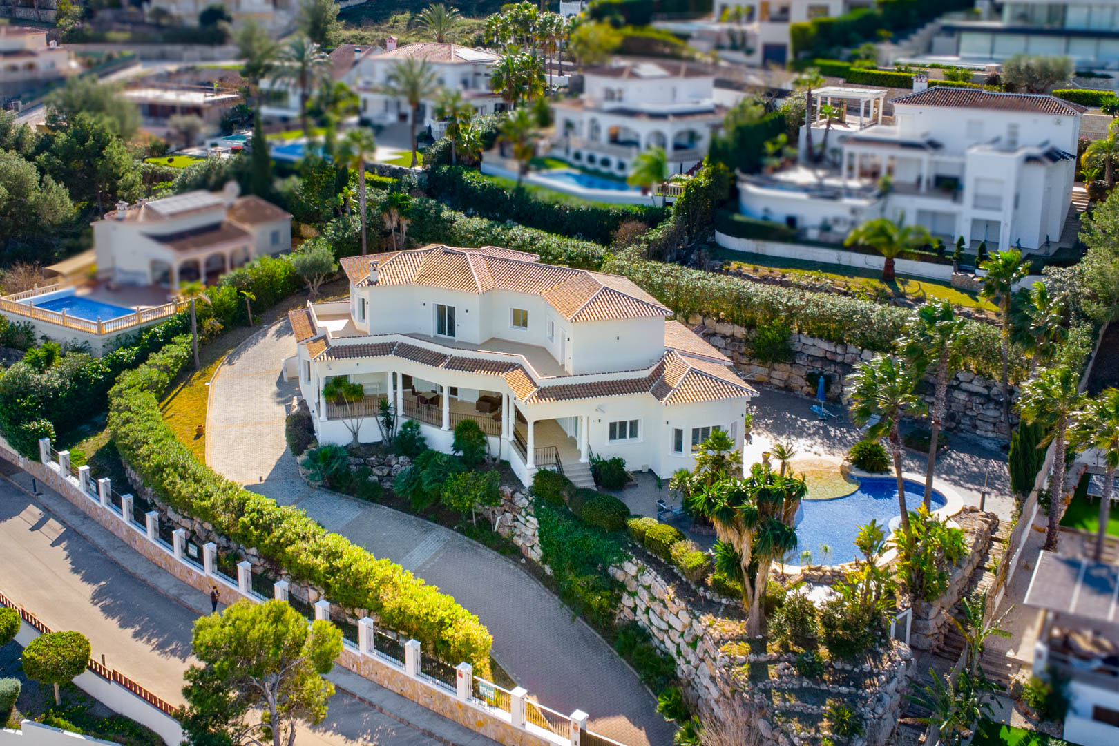 Vue aérienne de la villa aux façades blanches et toits de tuiles, révélant son allée pavée, sa piscine privée et ses jardins méditerranéens sur plusieurs niveaux.