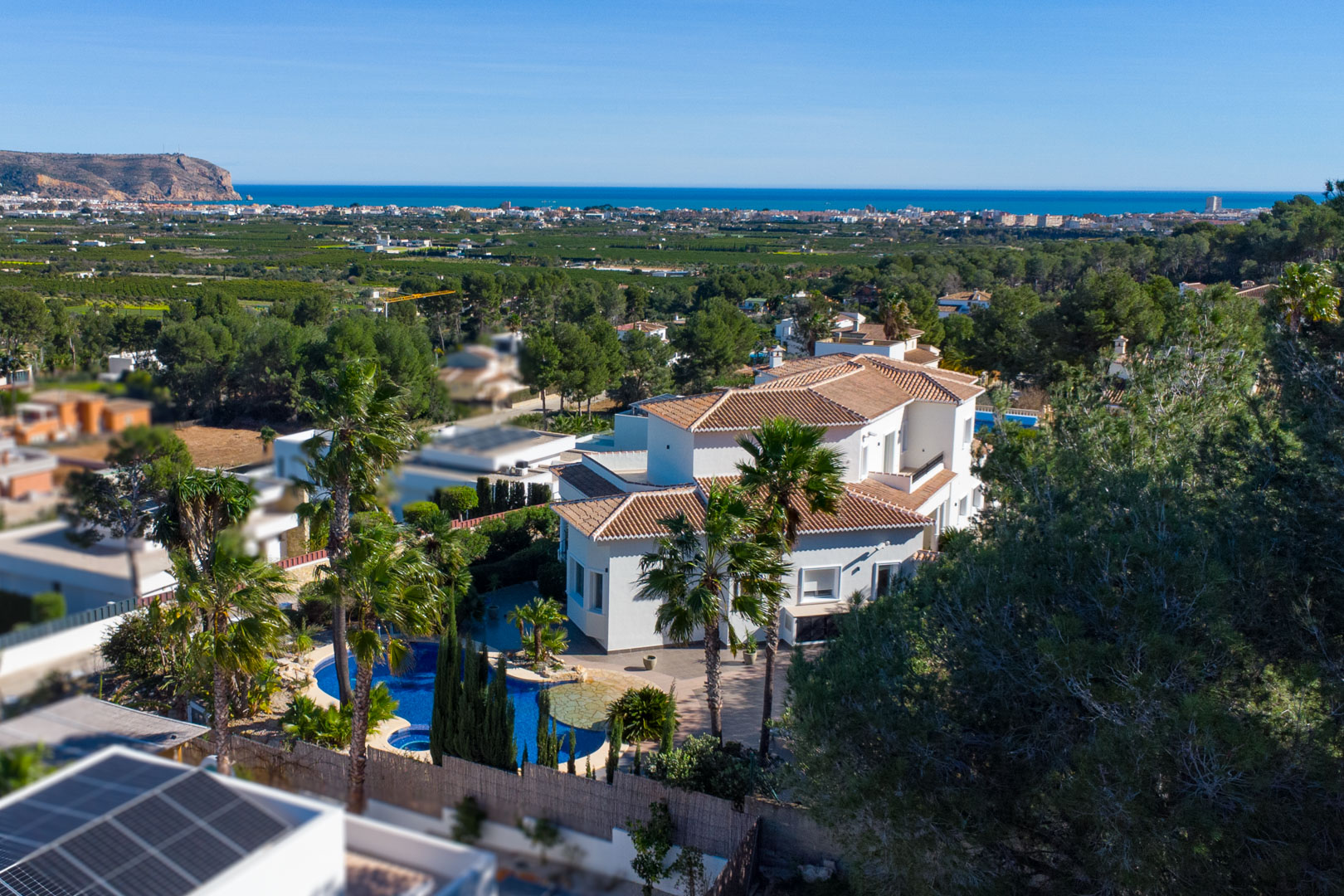Vue aérienne de la villa blanche aux toits de tuiles, incluant la piscine avec jacuzzi, le jardin méditerranéen et un panorama sur la côte et la mer.