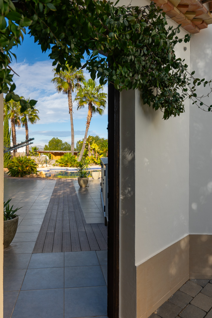Vue depuis le porche vers la piscine, avec un chemin en bois intégré à la terrasse carrelée et un jardin méditerranéen planté de palmiers.