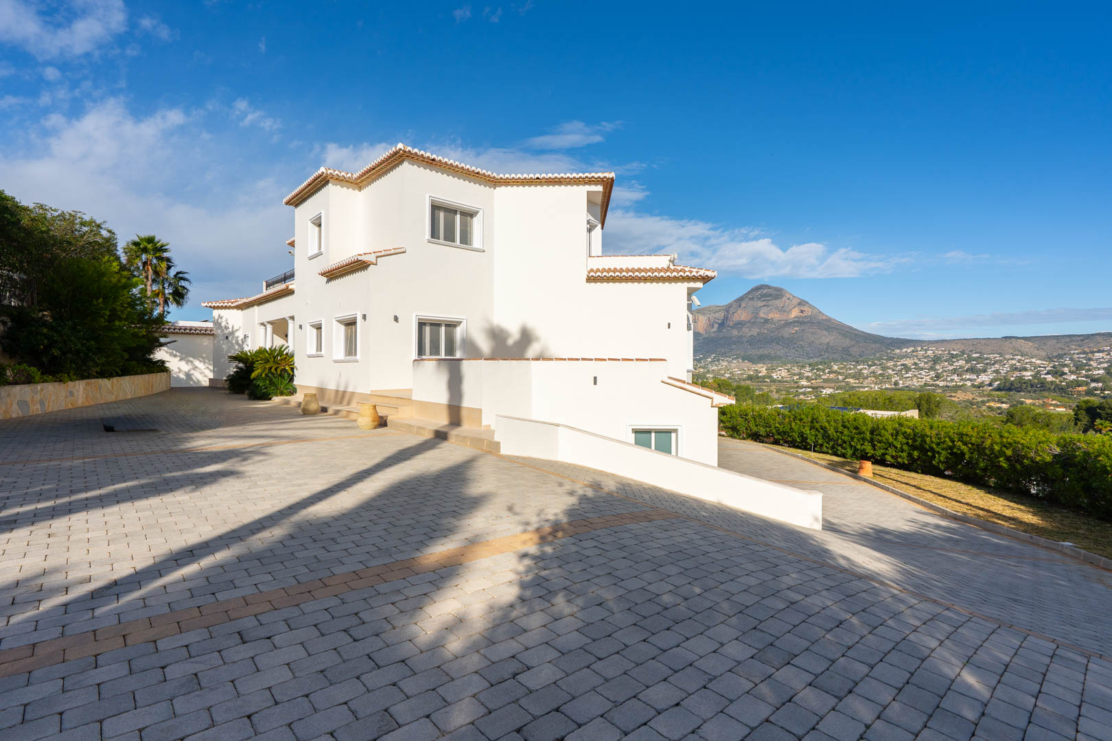 Vue extérieure de la villa à façade blanche, mettant en valeur la vaste allée pavée et la vue dégagée sur la montagne du Montgó.