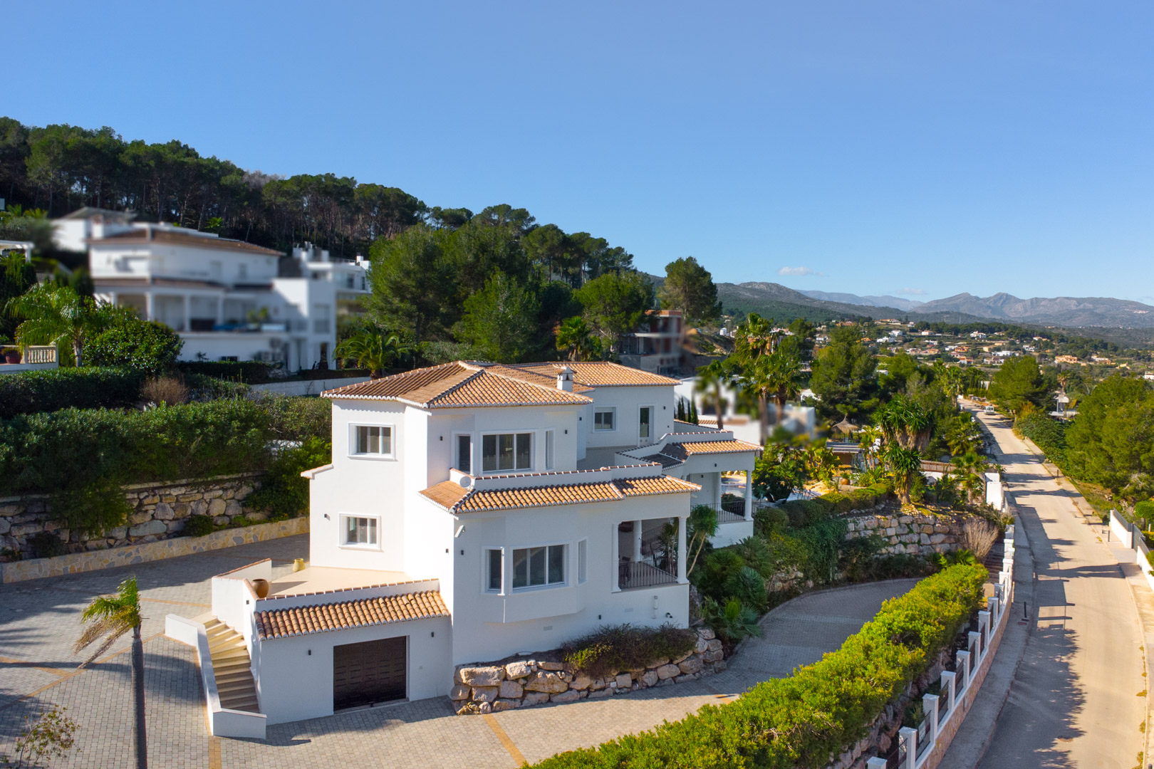 Vue extérieure de la villa aux façades blanches et toits en tuiles, illustrant ses différents niveaux, ses terrasses et son garage intégré dans un environnement arboré.