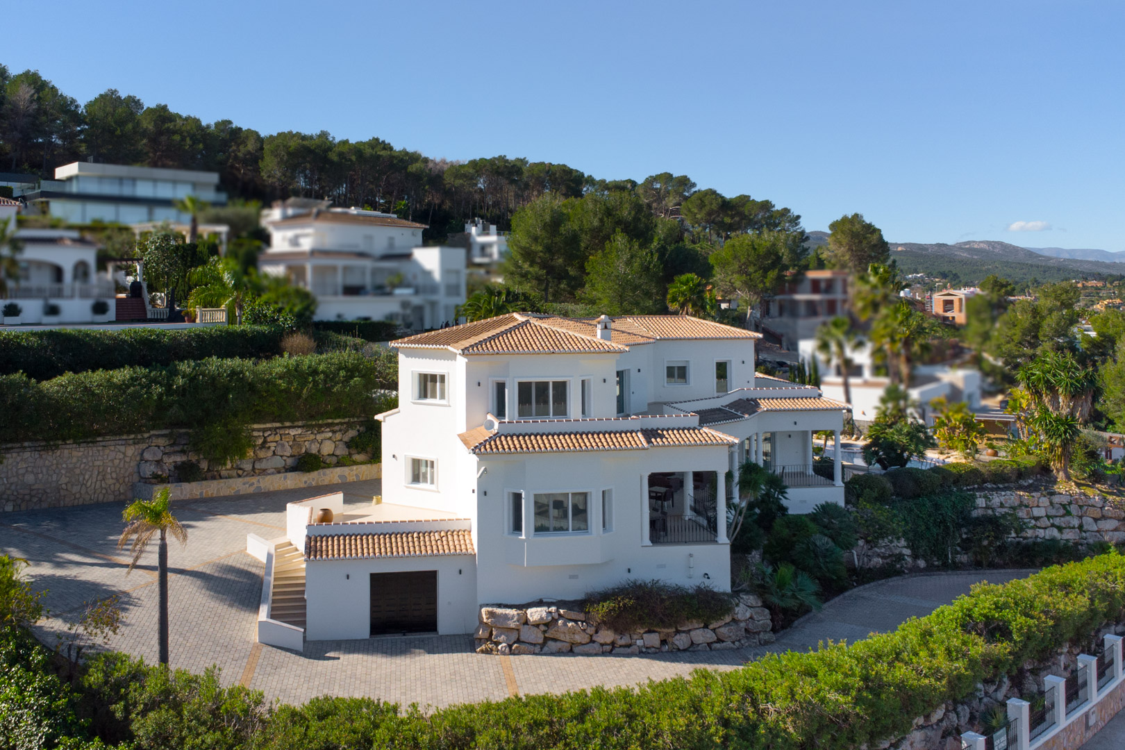 Vue extérieure de la villa méditerranéenne aux façades blanches, montrant les toits en tuiles, l'entrée pavée spacieuse et le garage en rez-de-chaussée.