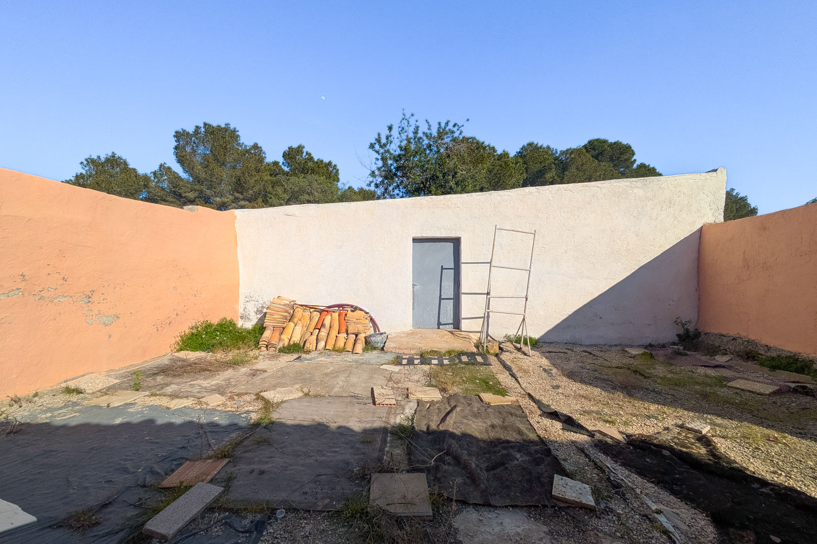 Cour extérieure fermée par de hauts murs, présentant une porte d'accès et des tuiles canal empilées sur un sol en cours de rénovation.