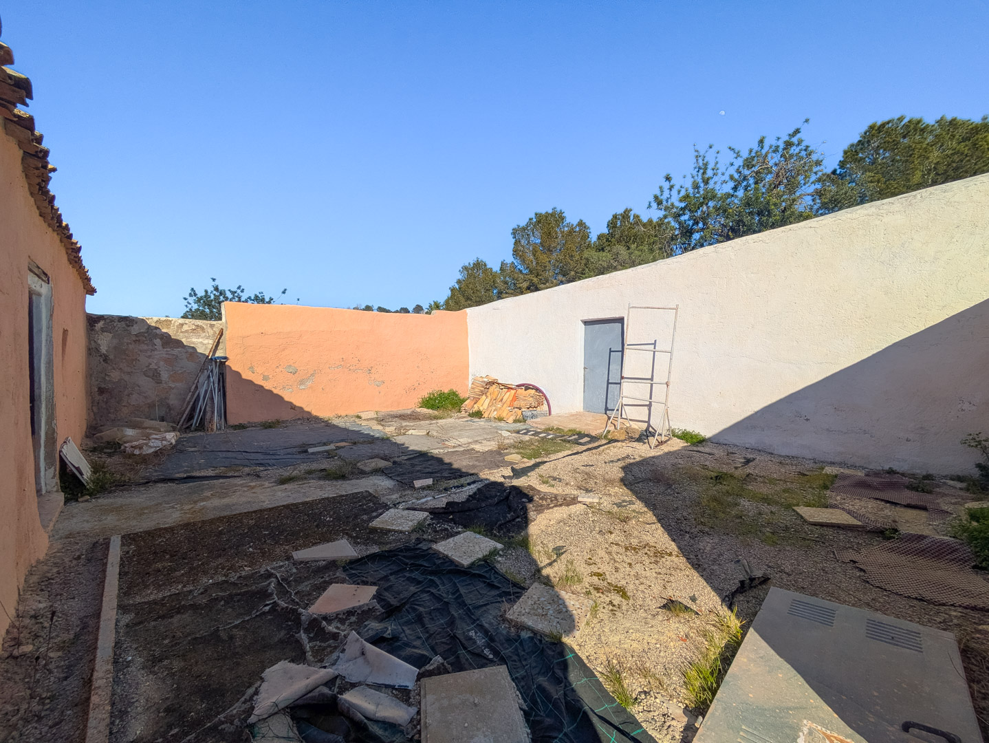 Cour intérieure fermée avec de hauts murs, une porte d'accès bleue et un sol brut avec bâches de protection, illustrant le potentiel de rénovation.