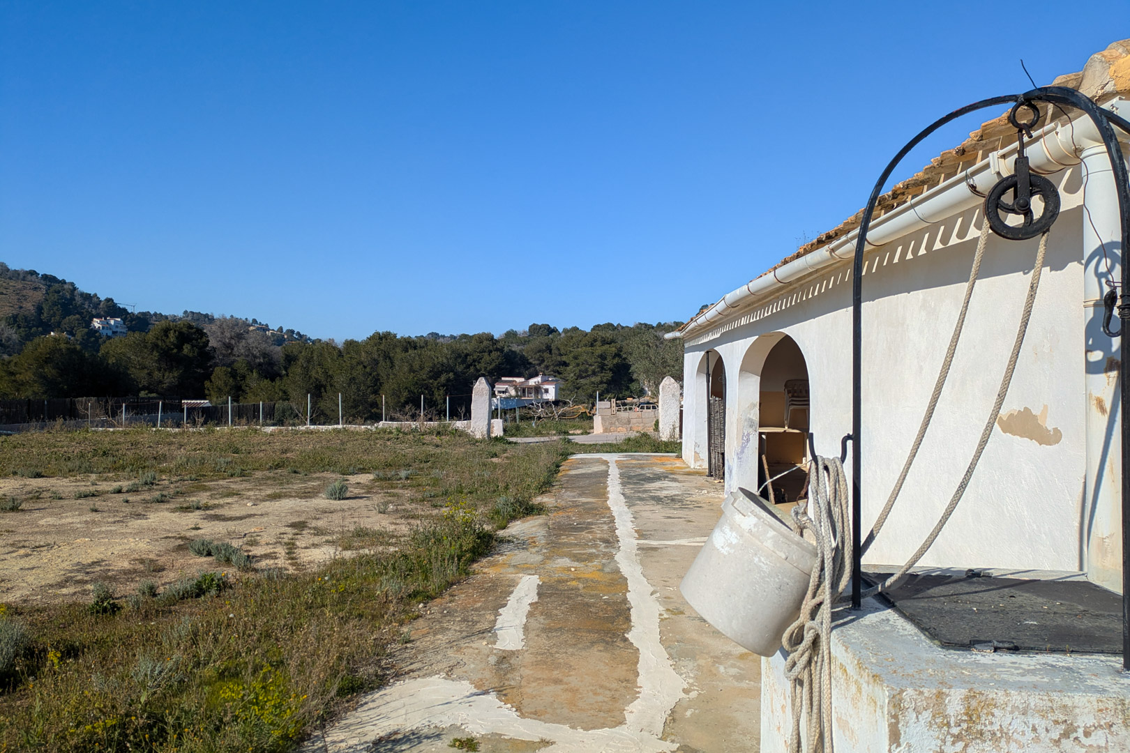 Extérieur de la finca avec un puits traditionnel et sa poulie, jouxtant la façade blanche à arches et un vaste terrain rustique plat en pleine nature.