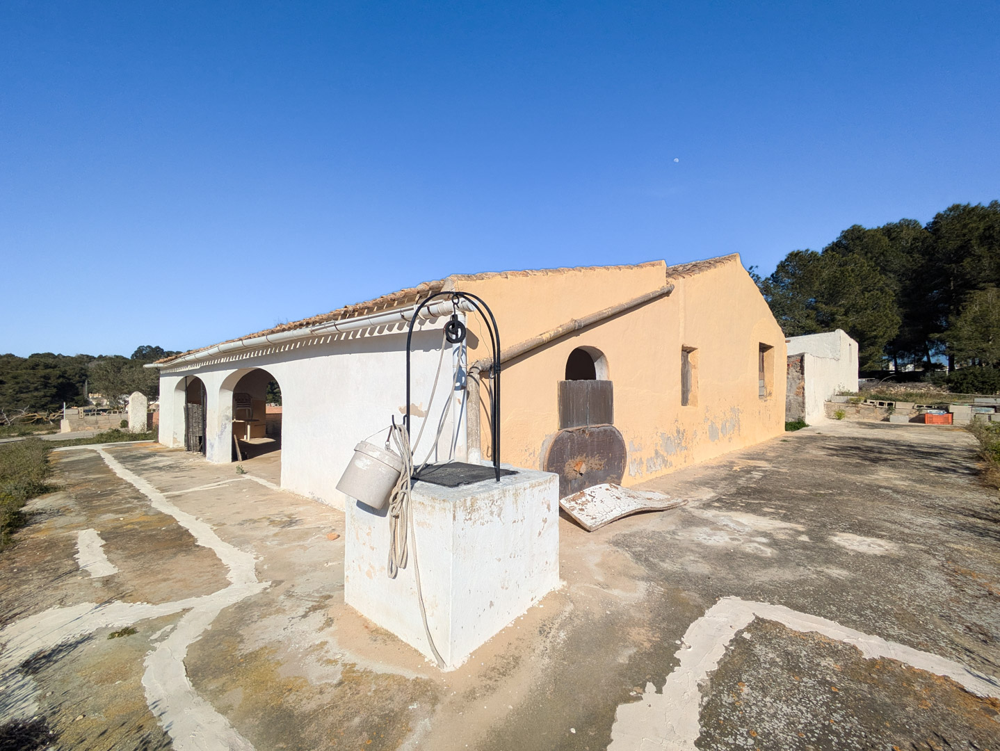 Façade de la finca traditionnelle avec porche à arcades, puits ancien au premier plan et vaste terrasse en pierre sous un ciel bleu.