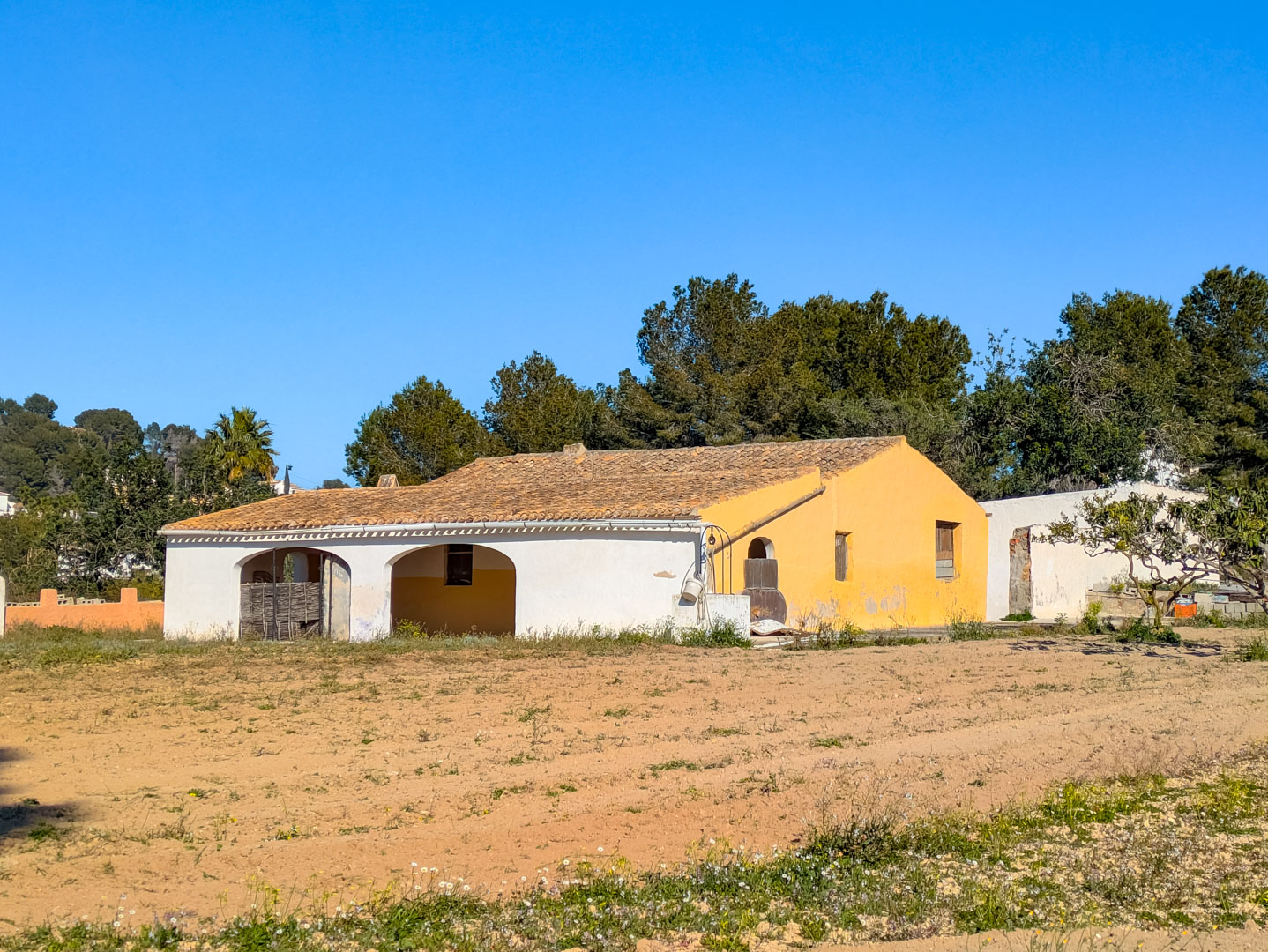 Façade de la finca traditionnelle avec porches voûtés et toit en tuiles, située sur un vaste terrain rustique entouré de pins méditerranéens.