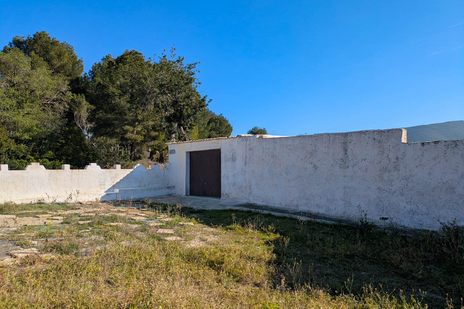 Mur de clôture blanc avec porte d'accès sombre, situé sur une parcelle rustique entourée de pins et d'une végétation méditerranéenne dense.