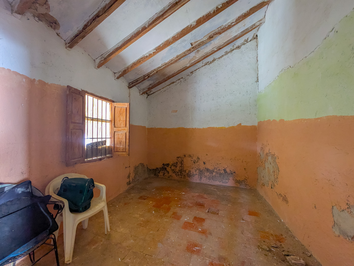 Pièce intérieure avec plafond incliné et poutres en bois apparentes, sol en terre cuite traditionnelle et fenêtre avec volets en bois d'origine.