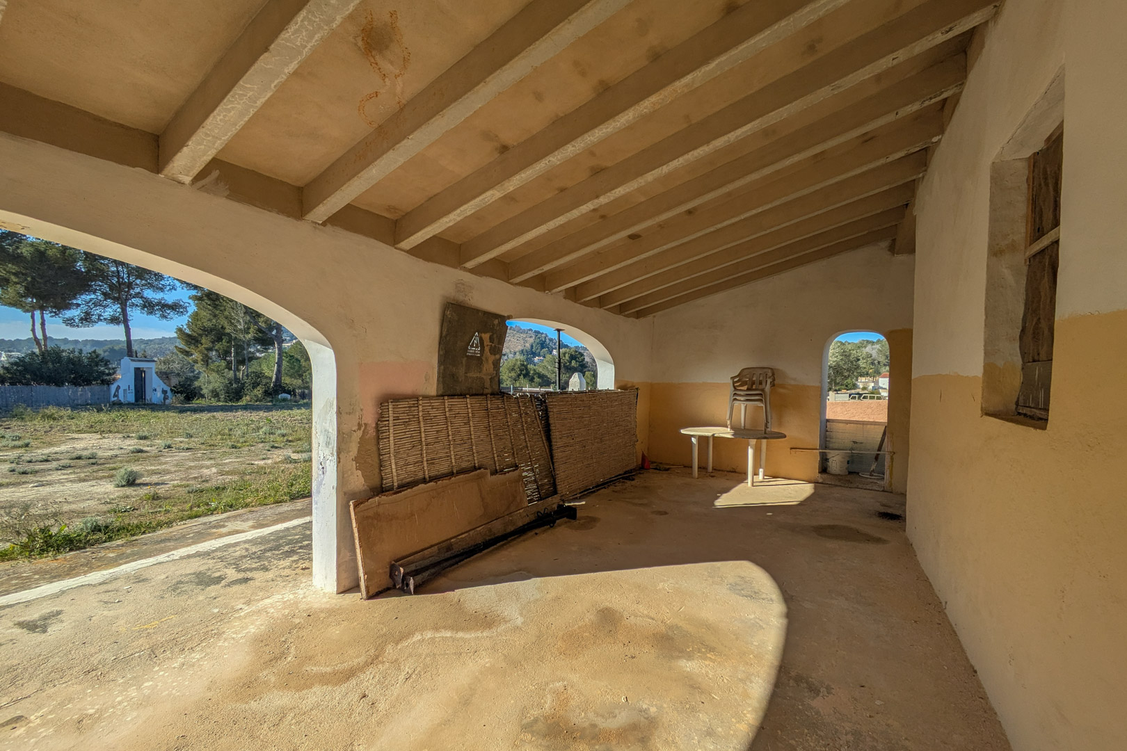 Porche couvert traditionnel avec poutres apparentes et ouvertures en arche, offrant une vue sur le terrain rustique et la nature environnante.