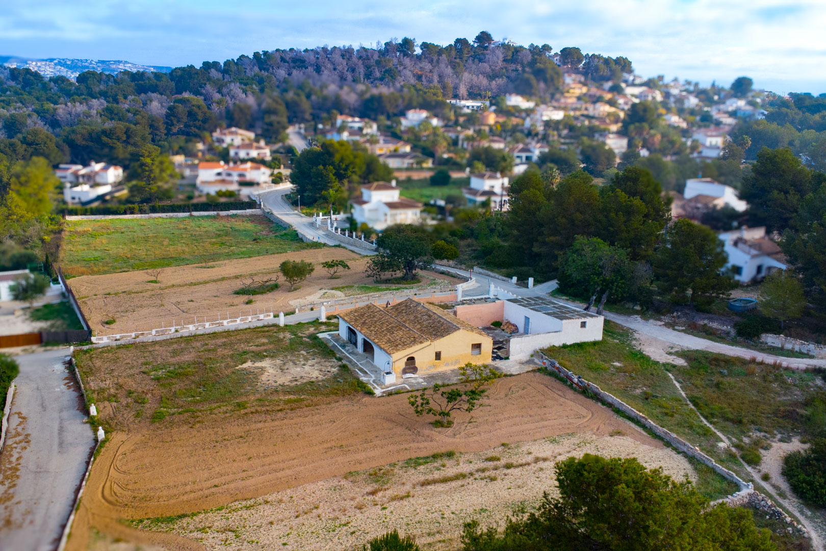 Vue aérienne de la finca traditionnelle avec toit en tuiles et façade ocre, implantée sur un vaste terrain rustique entouré de végétation et de villas.