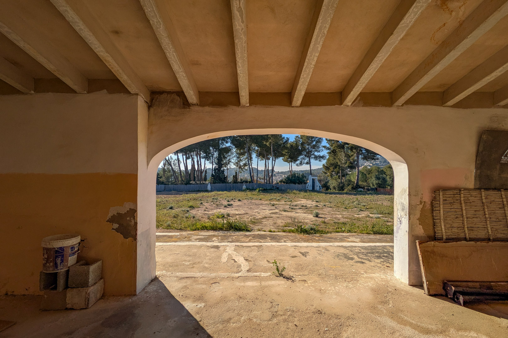 Vue depuis un porche voûté avec plafond à poutres, donnant sur le vaste terrain rustique et une haie de pins matures en arrière-plan.