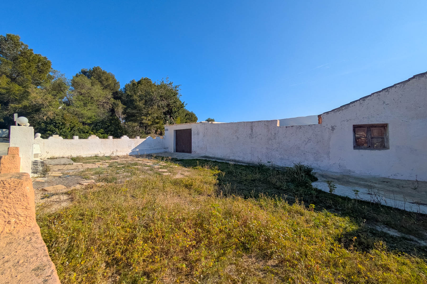 Vue extérieure de la façade blanchie à la chaux avec menuiseries en bois d'origine et cour rustique murée, entourée de pins méditerranéens.