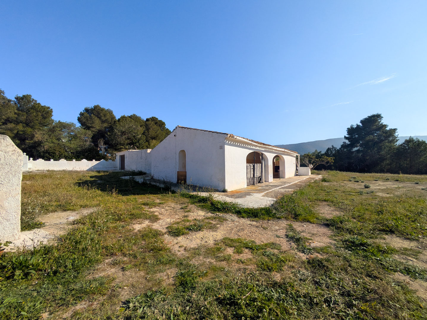 Vue extérieure de la finca traditionnelle aux murs blancs et porches voûtés, située sur un grand terrain rustique entouré de pins méditerranéens.