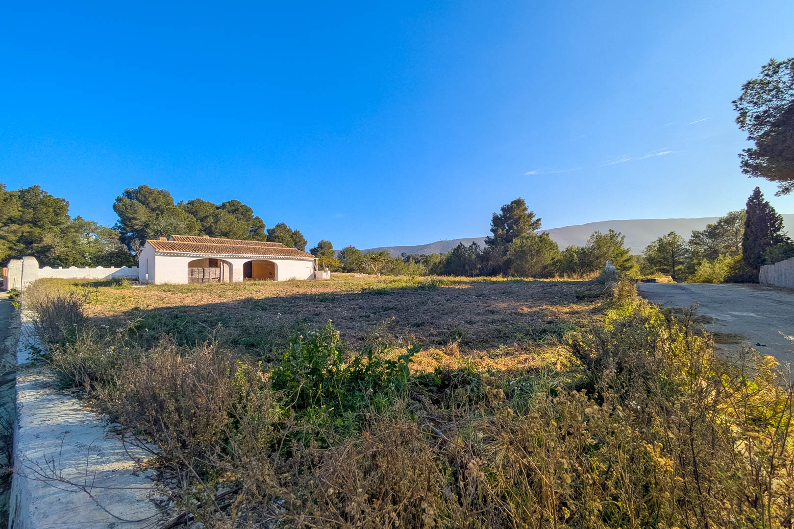 Vue extérieure de la finca traditionnelle aux murs blancs et porches voûtés, située sur un vaste terrain rustique entouré de pins avec vue sur les montagnes.