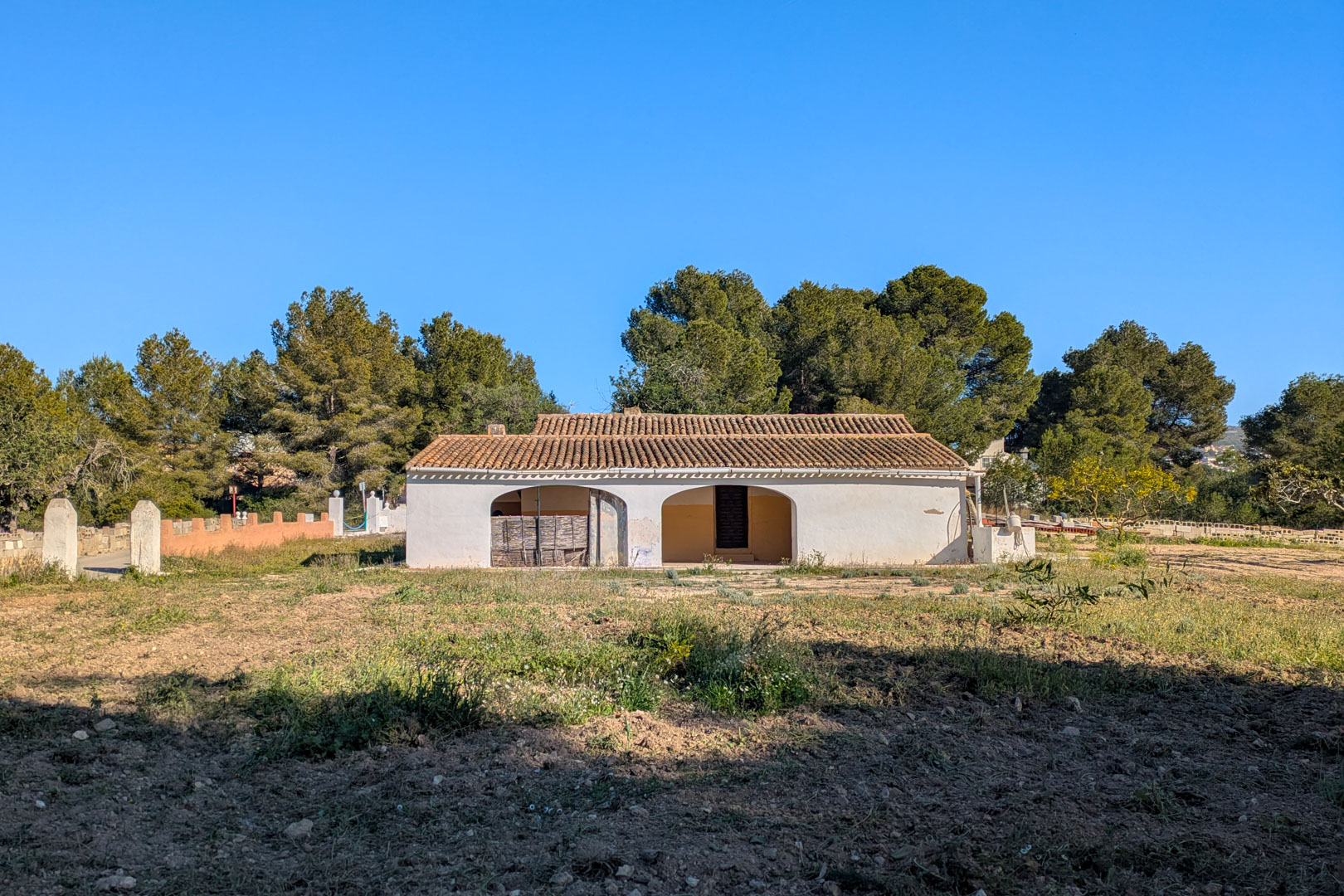 Vue extérieure de la finca traditionnelle avec façade blanche, porche à arches et toit en tuiles, située sur un terrain rustique entouré de pins.