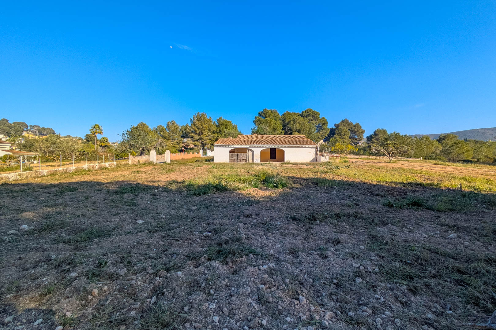 Vue extérieure de la finca traditionnelle avec porche à arcades et toit en tuiles, située sur un vaste terrain plat entouré de pins.