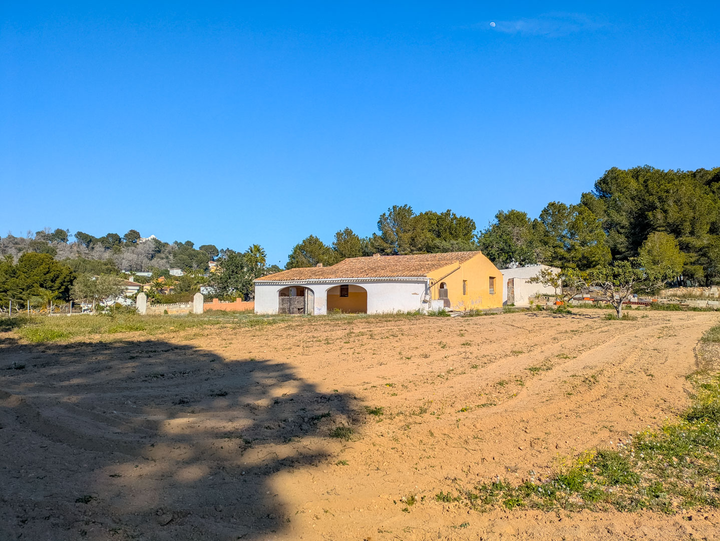 Vue extérieure de la finca traditionnelle avec son porche à arches, sa toiture en tuiles romanes et son vaste terrain rustique plat entouré de pins.