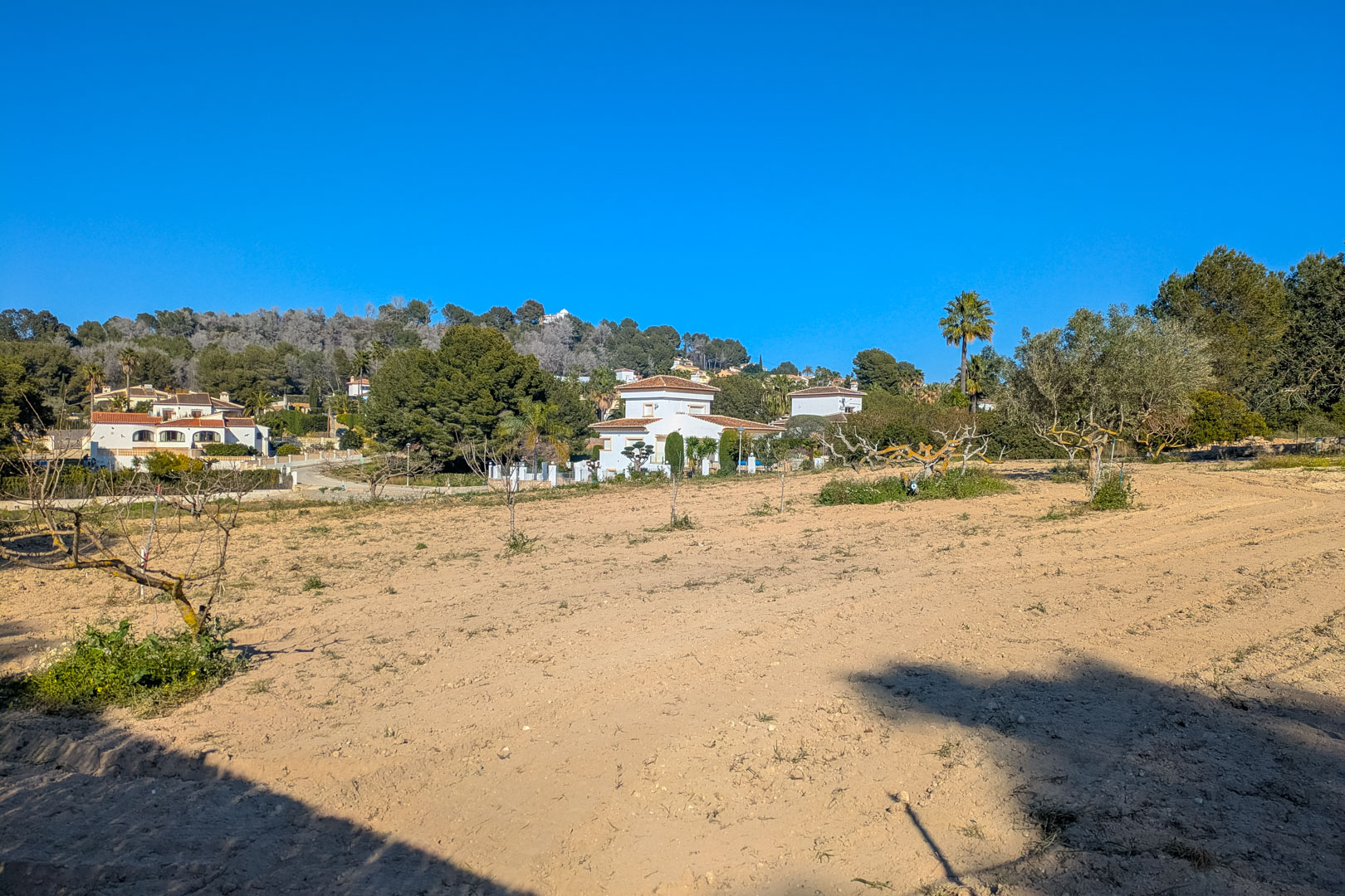 Vue panoramique de la vaste parcelle rustique plate, entourée de végétation méditerranéenne avec une vue dégagée sur les collines et les villas voisines.