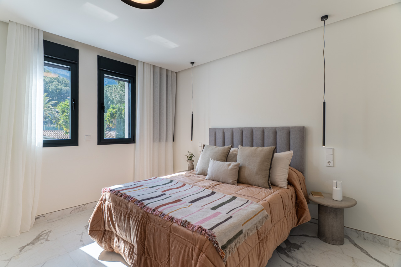 Chambre double avec sol en grès cérame effet marbre, tête de lit rembourrée grise et fenêtres à cadre noir offrant une vue sur la végétation environnante.