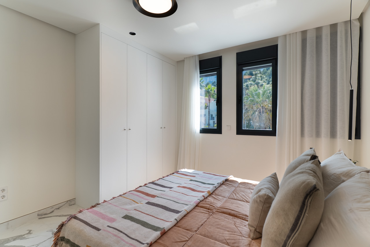 Chambre lumineuse avec placards intégrés blancs, sol en marbre et fenêtres offrant une vue sur la végétation environnante.