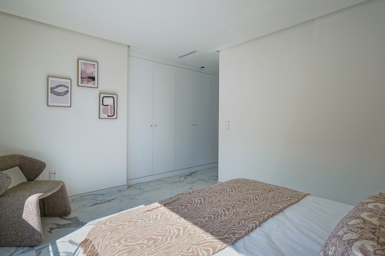 Chambre lumineuse avec placards intégrés blancs, sol en marbre veiné et un coin détente avec fauteuil design et cadres minimalistes.