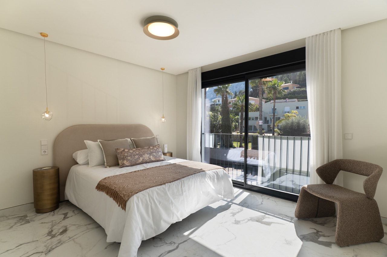 Chambre principale avec sol en grès cérame effet marbre et grandes baies vitrées coulissantes ouvrant sur un balcon privé avec vue dégagée.
