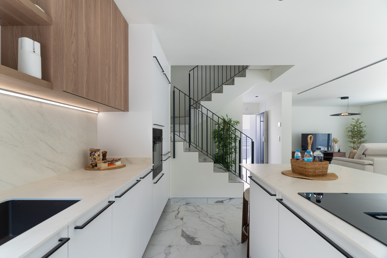 Cuisine à aire ouverte avec meubles blancs, plans de travail effet marbre et rangements en bois avec éclairage LED. On aperçoit l'escalier moderne et le salon en arrière-plan.