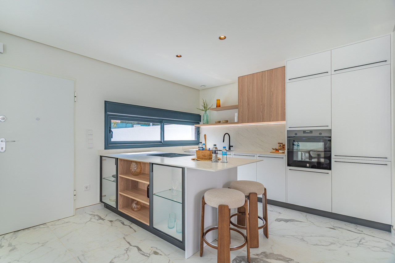 Cuisine moderne à aire ouverte avec îlot central, armoires blanches et touches de bois. Elle comprend des appareils intégrés et un sol en grès cérame effet marbre.