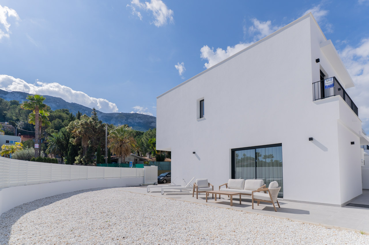 Façade blanche minimaliste avec terrasse pavée, mobilier d'extérieur et jardin en gravier blanc, offrant une vue sur le massif du Montgó.