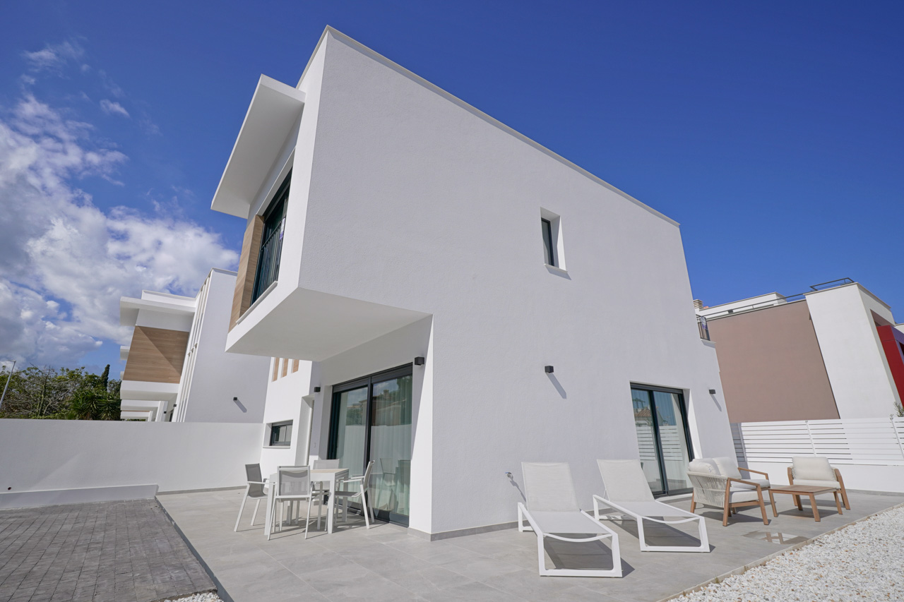Façade extérieure au design contemporain avec volumes blancs, terrasse dallée équipée de mobilier de jardin et espace de stationnement privé.