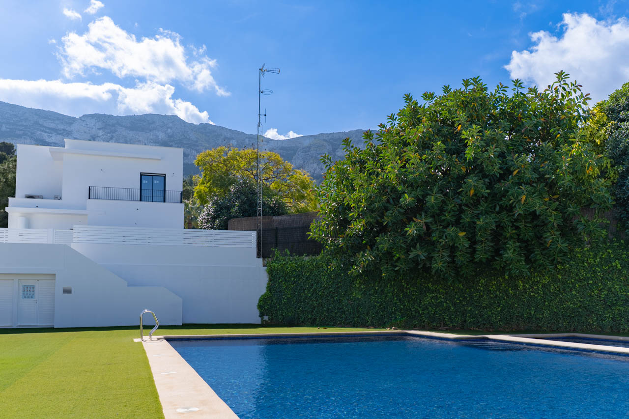 Piscine rectangulaire avec bordure en pierre et pelouse, située devant une façade blanche minimaliste avec le massif du Montgó en arrière-plan.